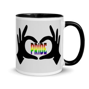 Puede incluir: Taza de café blanca con borde y asa negros. La taza presenta una silueta negra de dos manos haciendo un corazón con la palabra "PRIDE" en colores arcoíris dentro del corazón.
