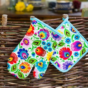 Può includere: Due guanti da forno colorati con un motivo floreale in tonalità di rosso, blu, verde, giallo e bianco. I guanti hanno un bordo blu chiaro e sono appesi a una recinzione di vimini intrecciato.