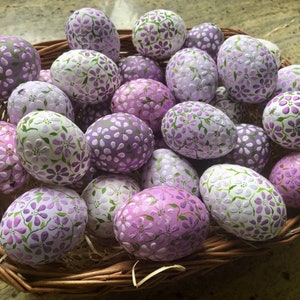 Può includere: Un cesto di vimini pieno di uova di Pasqua viola e bianche decorate con un motivo floreale.