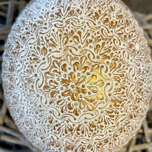Könnte beinhalten: Dekoratives, kugelförmiges Objekt mit einem weißen und goldenen Blumenmuster. Das filigrane Design bedeckt die gesamte Oberfläche und erzeugt ein strukturiertes Aussehen. Das Objekt ruht auf einem gewebten, naturfarbenen Untergrund.