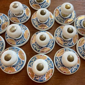 May include: A collection of decorative cabinet knobs. Each knob features a white, rounded handle with a brass-colored base. The circular base has a blue and gold floral design. The knobs are arranged on a wooden surface.