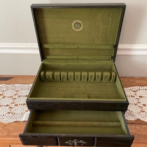 May include: An open, dark wooden silverware chest with a green felt interior. The chest has a top section with slots for utensils and a bottom drawer. A small, round gold emblem is visible inside the lid.
