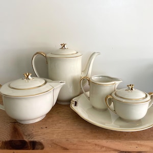 May include: A cream-colored tea set with gold trim. The set includes a teapot, a creamer, a sugar bowl with a lid, and a serving tray. The teapot and sugar bowl have decorative finials on their lids.