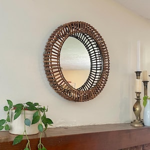 May include: A round mirror with a woven rattan frame. The mirror is hanging on a white wall above a wooden shelf. The mirror reflects the room. A potted plant and candlesticks are also visible.