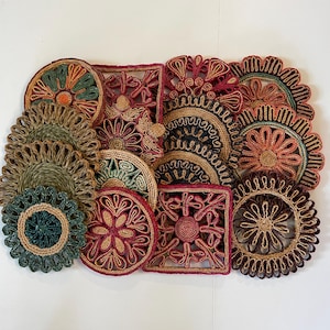 May include: A collection of decorative jute coasters in various shapes, including round and square. The coasters feature intricate floral and geometric designs in shades of green, red, brown, and natural jute tones. These coasters are ideal for protecting surfaces from drink rings.