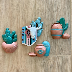 May include: A set of four decorative wall plaques featuring desert-themed motifs. The collection includes a green cactus in a peach pot, a white steer skull on a patterned textile, a terracotta vase with a green cactus, and two small clay-colored vessels.