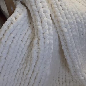 May include: A chunky knit blanket made of thick, soft white yarn. The textile features a consistent, repeating loop pattern created with large stitches, resulting in a textured, plush surface that drapes in soft folds across the frame.