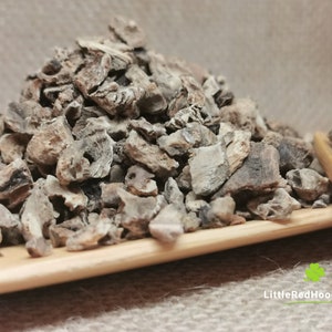 May include: A close-up of a pile of dried, brown, and grey burdock root pieces on a wooden spoon. The root pieces are irregularly shaped and have a rough texture.