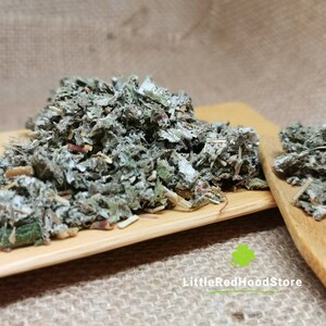May include: Dried green herbs, with a textured appearance, on a small wooden tray and spoon. The background is a neutral burlap fabric. The text "LittleRedHoodStore" is visible. The herbs appear to be leafy.
