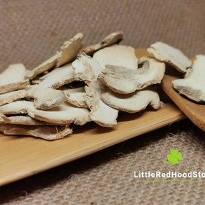 May include: Dried ginger slices, pale beige, are piled on a small, light-coloured wooden tray. The background is a textured, brown surface. The text "LittleRedHoodStore" is visible.