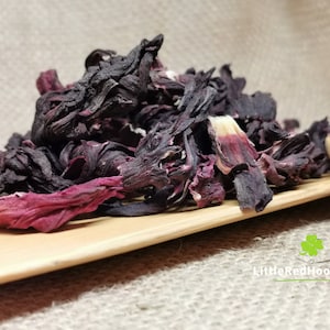 May include: A close-up of dried hibiscus flowers, displaying deep red and dark purple colours. The textured petals are layered, presented on a wooden surface. The image suggests a focus on natural ingredients, possibly for tea or culinary use.