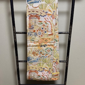 May include: A long, rectangular textile featuring a cream-colored base with intricate, multi-colored woven patterns of pine needles, floral motifs, and architectural elements, draped over a black, gold-accented wooden display stand.