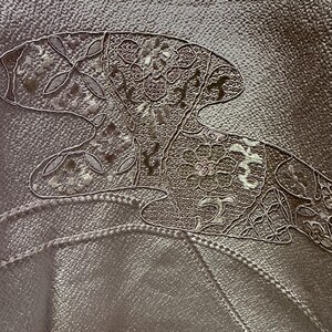 K&ouml;nnte beinhalten: Nahaufnahme eines hellbraunen Stoffes mit einem wei&szlig;en gestickten Design einer stilisierten Blume mit Bl&auml;ttern. Die Stickerei ist detailliert und kunstvoll.