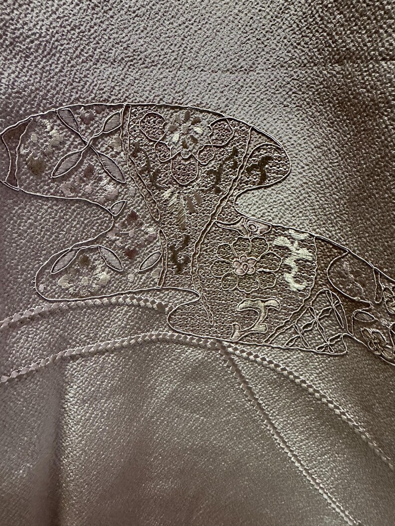 K&ouml;nnte beinhalten: Nahaufnahme eines hellbraunen Stoffes mit einem wei&szlig;en gestickten Design einer stilisierten Blume mit Bl&auml;ttern. Die Stickerei ist detailliert und kunstvoll.