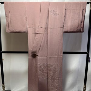 K&ouml;nnte beinhalten: Ein blasses rosa Kimono mit zarten gestickten Blumenmustern. Der Kimono h&auml;ngt an einem schwarzen Holzb&uuml;gel.