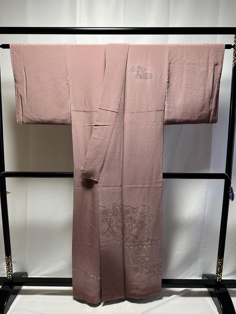 K&ouml;nnte beinhalten: Ein blasses rosa Kimono mit zarten gestickten Blumenmustern. Der Kimono h&auml;ngt an einem schwarzen Holzb&uuml;gel.