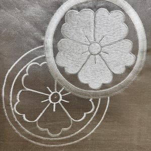Puede incluir: Primer plano de dos diseños florales bordados en hilo blanco sobre una tela gris plateada. Cada diseño presenta una flor dentro de un marco circular. El bordado tiene una apariencia detallada y texturizada.