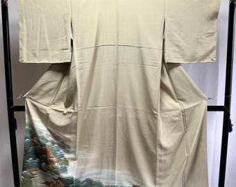 May include: A beige kimono displayed on a black stand. The kimono features a landscape design with houses, trees, and mountains in shades of green, brown, and blue at the bottom. The kimono is made of a smooth fabric.