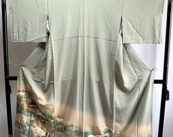 May include: A sage green kimono featuring a landscape scene along the bottom hem. The design depicts a forest with trees, small buildings, and white patches of snow, transitioning into a soft beige gradient against the solid light green fabric.
