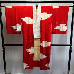 Könnte beinhalten: Rotes Kimono mit weißen Wolkenmustern, präsentiert auf einem schwarzen Gestell. Das Kimono hat einen weißen Kragen und einen beigen Schärpe. Der Stoff hat eine strukturierte Optik, und die Wolkenmuster sind gleichmäßig über dem Kleidungsstück verteilt.