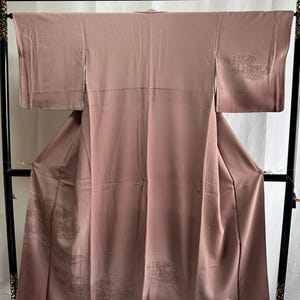K&ouml;nnte beinhalten: Ein mauvefarbener Seidenkimono mit gestickten Blumenmustern. Der Kimono h&auml;ngt an einem schwarzen Holzb&uuml;gel.