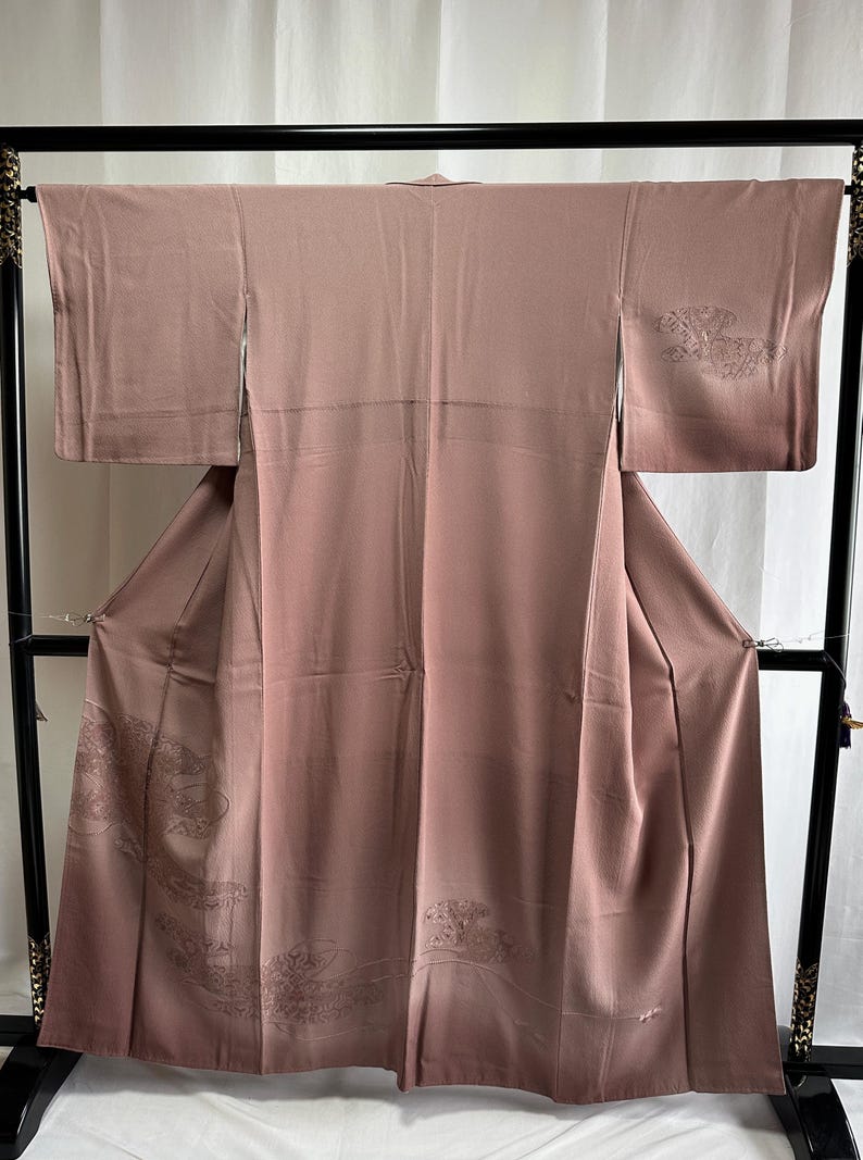 K&ouml;nnte beinhalten: Ein mauvefarbener Seidenkimono mit gestickten Blumenmustern. Der Kimono h&auml;ngt an einem schwarzen Holzb&uuml;gel.