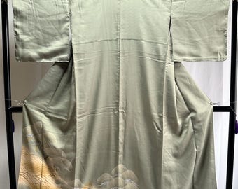 May include: A sage green kimono with a landscape design featuring stylized trees and water in shades of gold and beige. The traditional Japanese garment is displayed on a black stand, showcasing its flowing fabric and intricate details.
