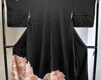 May include: A black kimono with a floral and fan design in shades of orange, peach, and white at the hem. The kimono is displayed on a black rack. Small white floral accents are on the shoulders.
