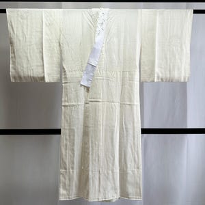 May include: A cream-coloured kimono hangs from a black bar. The kimono features a white collar with a floral design and a white sash. The fabric has a subtle textured pattern. The sleeves are wide and rectangular.