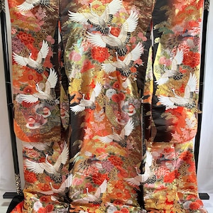 Könnte beinhalten: Ein formeller Kimono mit einem lebendigen Muster aus fliegenden weißen Kranichen, roten und orangefarbenen Blumenmotiven sowie goldenen Akzenten auf dunkelbraunem Grund. Das Kleidungsstück zeigt aufwendige Stickereien und einen dicken, gepolsterten Saum aus leuchtend rotem Stoff.
