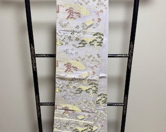 Op de afbeelding: Een zwarte kimonorek met gouden accenten houdt een lichtgrijze kimono vast. De kimono heeft een gedetailleerd patroon met gouden, groene en rode elementen die een landschap uitbeelden. De rek heeft een decoratieve basis.