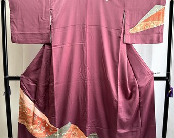 May include: A mauve-colored kimono displayed on a black stand. The kimono features decorative patterns in shades of orange, gray, and beige along the bottom and sleeves. A white triangle is sewn on the upper right side. The garment is made of a smooth, silky fabric.