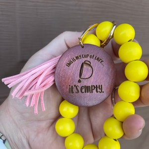 Può includere: Un portachiavi con perline gialle e un ciondolo in legno che dice "This is my cup of care... it's empty". Il ciondolo ha una piccola immagine di una tazza di caffè con una goccia di caffè che ne esce. Il portachiavi ha un fiocco rosa.