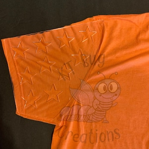 May include: Orange t-shirt with a clear plastic stencil of stars on the sleeve. The stencil is positioned over the upper part of the sleeve. The t-shirt has the text "Kit Bug Creations" printed on the bottom right corner.
