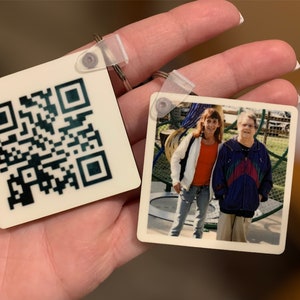 Puede incluir: Dos llaveros cuadrados con un anillo de plástico transparente. Un llavero tiene un código QR en blanco y negro. El otro llavero tiene una fotografía en color de dos personas de pie en el exterior.