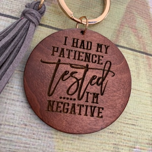 May include: A wooden keychain with a gray tassel. The keychain has the text "I had my patience tested... I'm negative" engraved on it.