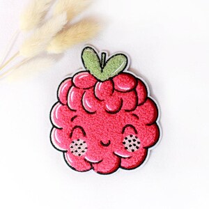 Könnte beinhalten: Ein himbeerförmiger Stickaufnäher mit einer leuchtend rosa strukturierten Oberfläche, einem kleinen grünen Stiel und einem lächelnden Gesicht mit zwei rosa Wangen, die mit schwarzen Punkten verziert sind. Der Aufnäher hat einen weißen Rand.