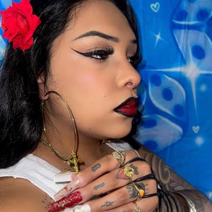 May include: A woman with dark hair, wearing a red rose, large gold hoop earrings, and multiple gold rings. Her nails are long and decorated with red and gold designs. She has dark lipstick and eyeliner, and a gold cross necklace.