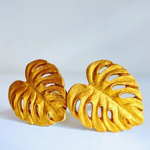 Könnte beinhalten: Ein Paar goldfarbene Ohrringe in Form von Monstera-Blättern. Die Ohrringe sind aus glänzendem, metallischem Material gefertigt und haben ein detailliertes Design.