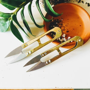 May include: Two gold-toned fabric scissors with sharp blades. The scissors have a unique design with a curved handle and a pointed tip.