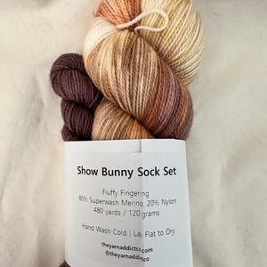 May include: A skein of brown, orange, and cream yarn with a label that reads "Show Bunny Sock Set Fluffy Fingering 80% Superwash Merino, 20% Nylon 480 yards / 120 grams Hand Wash Cold | Lay Flat to Dry theyarnaddictco.com @theyarnadditco"