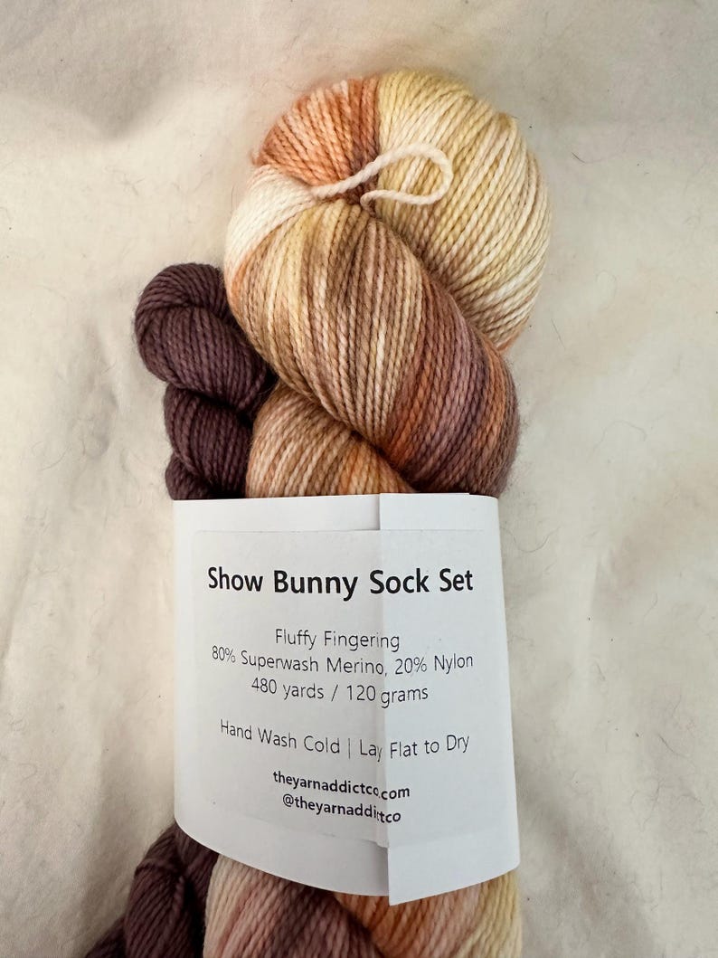 May include: A skein of brown, orange, and cream yarn with a label that reads "Show Bunny Sock Set Fluffy Fingering 80% Superwash Merino, 20% Nylon 480 yards / 120 grams Hand Wash Cold | Lay Flat to Dry theyarnaddictco.com @theyarnadditco"
