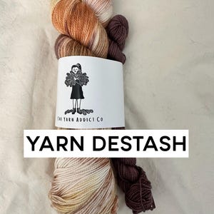 May include: Two skeins of yarn, one in a multi-colored ombre pattern of brown, orange, and cream, and one in a solid dark brown. The yarn is wrapped around a white label with the text "The Yarn Addict Co." and the words "YARN DESTASH" in black text.