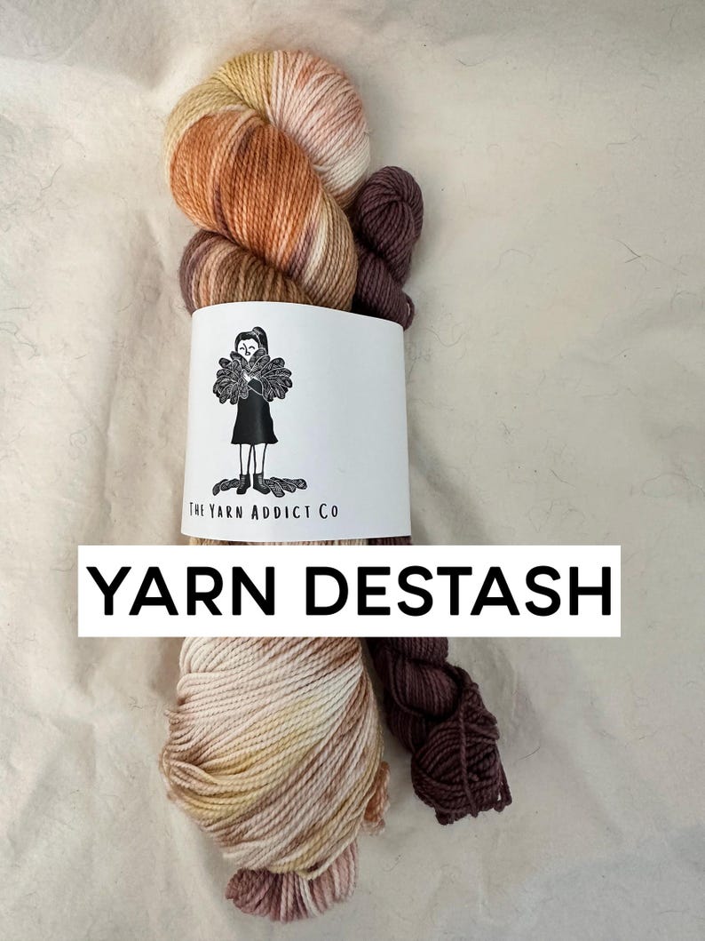 May include: Two skeins of yarn, one in a multi-colored ombre pattern of brown, orange, and cream, and one in a solid dark brown. The yarn is wrapped around a white label with the text "The Yarn Addict Co." and the words "YARN DESTASH" in black text.