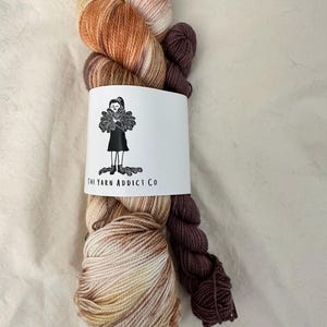 May include: Two skeins of yarn, one with a brown, orange, and white color gradient and the other a solid brown color. The yarn is wrapped around a white card with the text "The Yarn Addict Co."