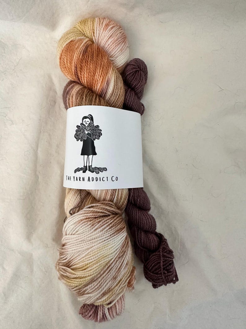 May include: Two skeins of yarn, one with a brown, orange, and white color gradient and the other a solid brown color. The yarn is wrapped around a white card with the text "The Yarn Addict Co."