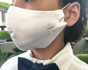 May include: A white satin face mask with a silver cross embroidery. The mask is being worn by a child wearing a white shirt and a dark blue bow tie.