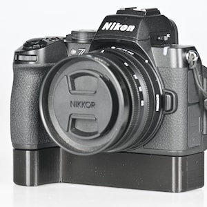 May include: A black Nikon camera with a lens cap that says "NIKKOR" on it. The camera has a black grip and a black battery grip.