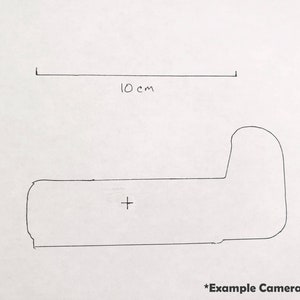 May include: A hand-drawn sketch of a camera, with a length of 10 cm indicated above the camera outline. The sketch includes a crosshair and the text "*Example Camera Sketch" at the bottom.