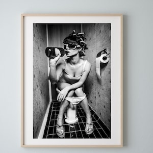 Puede incluir: Fotografía en blanco y negro de una mujer sentada en un inodoro, con gafas de sol y bebiendo de una botella. Lleva el pelo en rulos. Hay un rollo de papel higiénico en la pared detrás de ella.